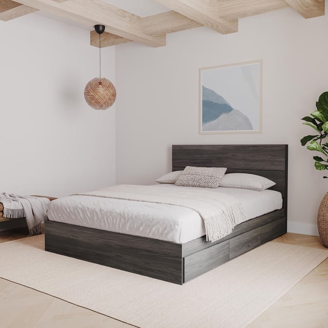 Nexera 2 Piece Bedroom Set with a bark grey storage bed and matching rustic plank-effect headboard, shown in a 3/4 front view. The bed includes three drawers on metal slides that can be installed on either side. The set is styled in a modern bedroom with light wood ceiling beams, a woven pendant light, neutral bedding, a beige rug, abstract wall art, and a large potted plant, ideal for combining storage and contemporary design.