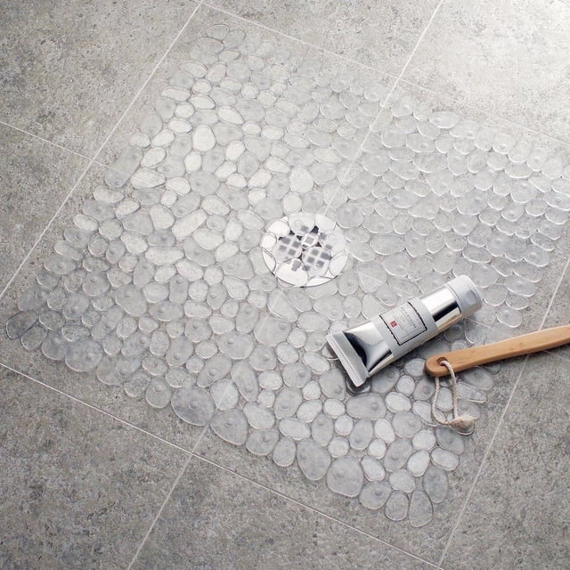 Tapis de Baignoire en PVC Format Étal de Galets avec Ventouses Transparentes en Forme de Galets et Trous de drainage sur un Plancher de Salle de Bain Gris à Carreaux, Offrant une Sécurité Antidérapante et une protection Anti-Moisissure, Exposé avec un tube de Produit de Bain Argenté et une Brosse de Bain en Bois.