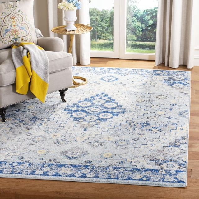 Safavieh Madison Sophie Geometric Area Rug with a blue and cream distressed geometric pattern, power loomed for durability, placed on hardwood flooring beside a beige armchair with a yellow and gray throw, near a gold leaf-shaped side table and beige curtains framing large windows.