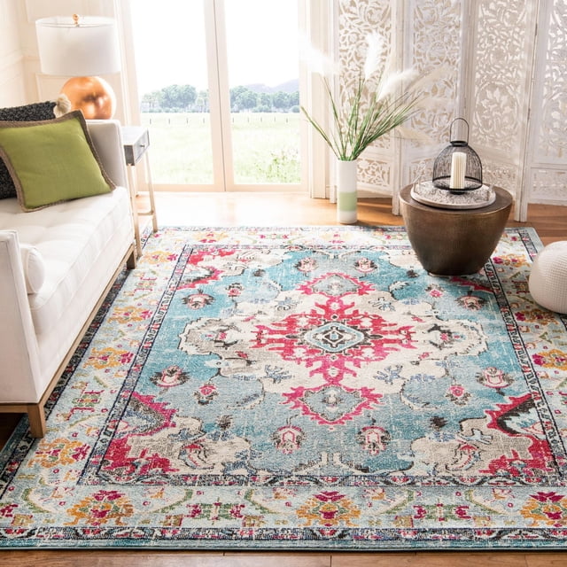 Safavieh Monaco Toria Traditional Area Rug with vibrant blue and pink Persian-inspired patterns in durable polypropylene with mixed loop pile, displayed in a bright living room featuring a white sofa with green cushion, wooden side table with copper lamp, decorative vase with pampas grass, and round brown coffee table with candle lantern. Suitable for adding Bohemian-chic flair to countryside or city homes.