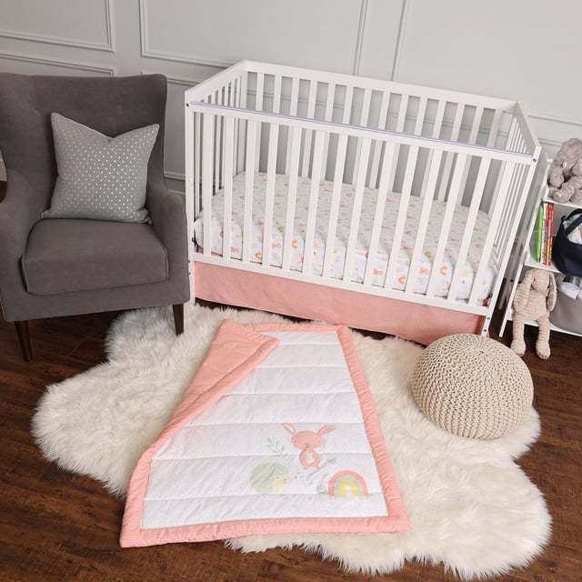 George Baby Ensemble de Literie de Bassinette 3 Pcs dans une Chambre de Bébé avec un Bassinette Blanc Présentant un Drap-Housse 100 % Coton Imprimé d'Icônes pastel et une Jupe de Bassinette Rose. la Couette avec une Bordure Rose Présente un Lapin, un Hérisson et un Motif Arc-en-Ciel, Posée sur un Tapis Duveteux Blanc à Côté d'Un pouf Tricoté beige et d'Un Fauteuil Gris avec un Coussin à Pois.