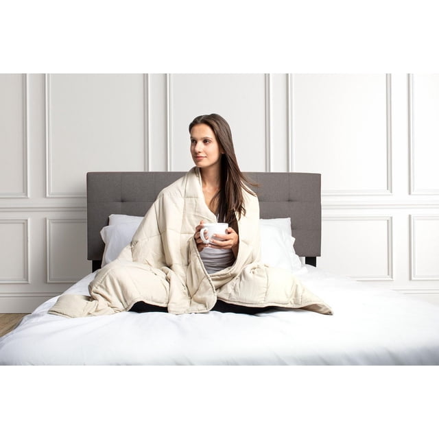 Safdie & Co. Weighted Blanket Taupe wrapped around a person sitting cross-legged on a white bed with a gray tufted headboard, holding a white mug, in a bright room with white paneled walls.