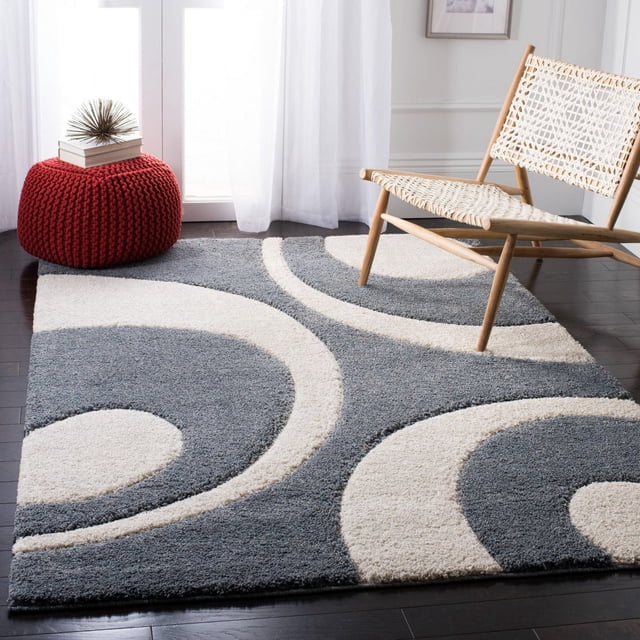 Safavieh Florida Mirabelle Contemporary Shag Area Rug featuring a plush high-low pile with grey and cream abstract swirl patterns on dark hardwood flooring, styled with a woven wooden chair and a red knitted pouf with books, suitable for enhancing texture and comfort in living areas.