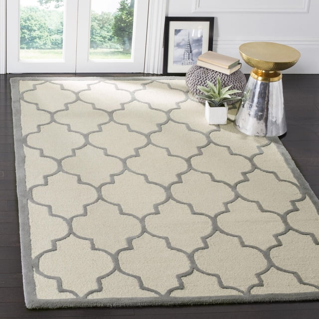 Safavieh Cambridge Kevin Geometric Area Rug in cream with grey trellis pattern and border, hand-tufted wool from the Cambridge Collection, displayed on dark hardwood floor with a silver and gold metal side table, grey knitted pouf with books, and a small potted succulent, suitable for adding modern American style and texture to home or office accent spaces.