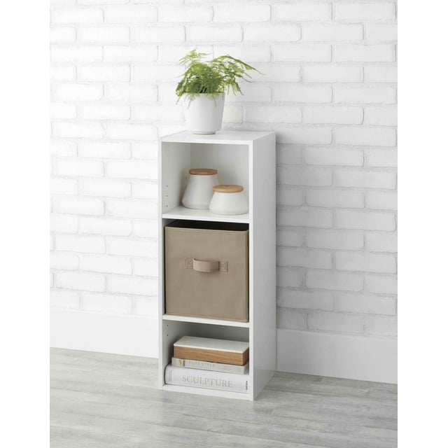 Mainstays Bibliothèque Organisatrice Réglable à 3 Tablettes en Fini Blanc avec Deux Tablettes Ajustables et Dos Fermé. l'Étagère Supérieure Contient Deux Pots en Céramique Blancs avec Couvercles en Bois, l'Étagère Centrale Présente un Bac de Rangement en Tissu beige, et l'Étagère Inférieure Contient Trois Livres Empilés. une Plante en Pot Verte s'Asseoit sur le Dessus contre un Mur de Briques Blanc et un Plancher de Bois Gris, Idéale pour le Rangement et l'Organisation de la Chambre des Enfants.