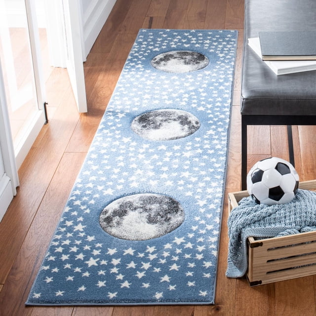 Safavieh Carousel Kids Moon in the Stars Area Rug, a rectangular blue kids' rug featuring three detailed moon phases and scattered white stars, power-loomed with durable synthetic yarns, placed on hardwood flooring beside a gray bench and a wooden crate with a soccer ball and knit blanket, ideal for kids' room decor.