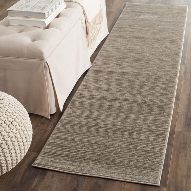 Safavieh Vision Adrasteia Overdyed Solid Area Rug in taupe with subtle horizontal ribbed texture, placed on dark hardwood floor beside a beige slipcovered sofa with a fringed throw and stacked books, next to a knitted pouf, ideal for contemporary living rooms or hallways.