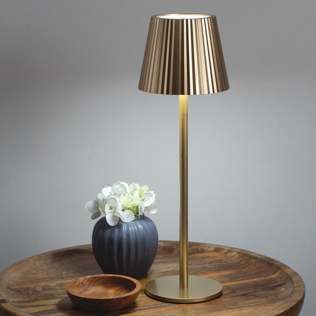 Merkury Innovations 14" Portable LED Metal Touch Lamp Gold with a fluted gold metal shade and slim cylindrical base, shown on a round wooden tray with a dark gray ribbed vase holding white flowers and a small wooden bowl, set against a plain gray wall. The lamp features touch controls and adjustable warm, cool, and natural white LED lighting, suitable for creating ambient lighting in any room.