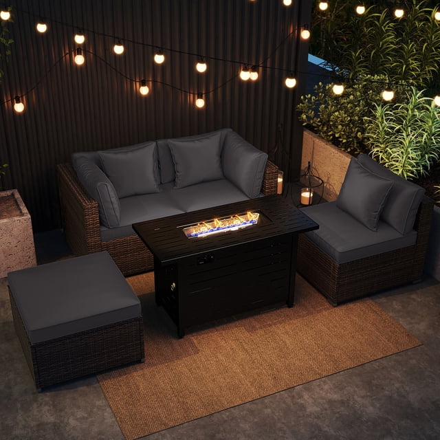 Costway 5 PCS Patio Furniture Set Heavy-duty Frame Rattan Sofa Set with 60,000 BTU Firepit Table in gray. The set includes two corner sofas, one armless sofa, an ottoman, and a firepit table with a black metal top. The furniture is crafted with premium PE rattan and features gray cushions with zippered covers for easy cleaning. The firepit table doubles as a coffee table when covered with its lid. The setup is displayed in an outdoor patio setting with string lights and greenery in the background.