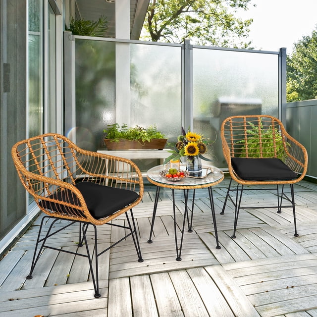 Costway 3PCS Patio Rattan Bistro Furniture Set Cushioned Chair Table Black. This 3-piece outdoor furniture set includes two rattan chairs with black cushioned seats and a round tempered glass coffee table. The chairs feature a wide backrest and curved armrests for ergonomic comfort, while the table provides a sturdy surface for drinks or decor. The set is constructed with weather-resistant rattan and a powder-coated steel frame for durability. Ideal for patios, balconies, or gardens, the set is shown in a modern outdoor setting with plants and sunlight.