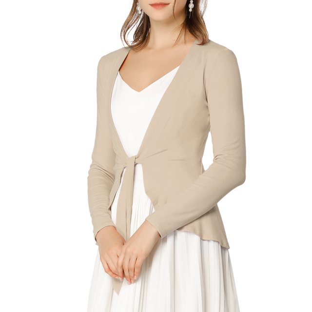 Allegra K Juniors Tie Open Front Long Sleeve Knit Metallic Ruffled Crop Cardigan in a solid beige colour. The cardigan features a tie-open front, ruffled hem, ribbed texture, and slim fit design. Made from a blend of polyester, rayon, and spandex, it is styled as a cropped cardigan with long sleeves. The model is wearing the cardigan over a white dress, showcasing its waist-defining cut and versatile design suitable for daily wear, office, or casual outings.