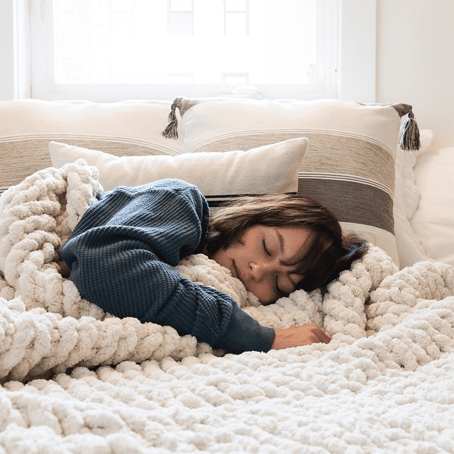 Comtest Chunky Knit Throw Blanket Chenille, Beige, 50"x60" shown in a cozy bedroom setting. The blanket, crafted from soft chenille yarn, features a chunky knit design in a neutral beige tone. A person is wrapped in the blanket, resting on a bed with decorative pillows in the background. Perfect for kids' bedding, warmth, and versatile use as a throw or gift.
