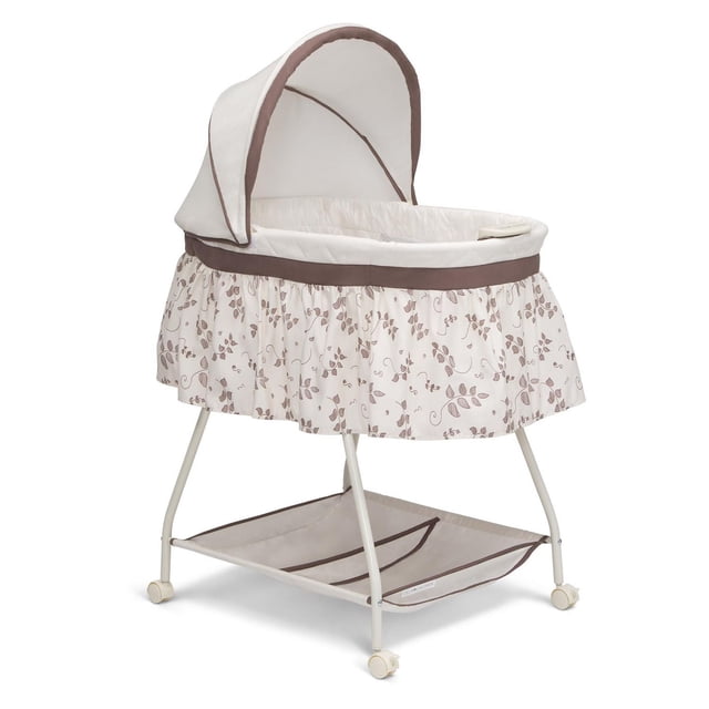 Delta Children Deluxe Sweet Beginnings Bassinet in Falling Leaves pattern with cream fabric and brown trim, featuring a removable canopy, ruffled skirt with leaf print, locking wheels for easy mobility, and a large storage basket underneath for baby essentials.