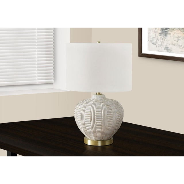 Monarch Specialties Lighting 21"h table lamp with a textured cream resin base featuring vertical ribbed detailing on a round gold metal footing, topped by an ivory linen fabric drum shade with a round gold finial, placed on a dark wood table beside a window with white blinds and beige walls, suitable for living room or bedroom use.