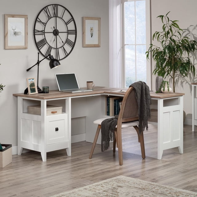 Sauder August Hill L-Desk in Soft White Finish with L-shaped wood-tone top and white base, featuring a file drawer with full extension slides for letter-size hanging files, cord management, and a lower shelf with books. The desk is set in a bright home office with a wooden chair draped with a gray throw, a black adjustable desk lamp, laptop, coffee mug, framed animal prints, a large black metal wall clock, and a green plant near a window with sheer curtains. Ideal for computer work and organizing office essentials, it is also suitable for crafting or study tasks.