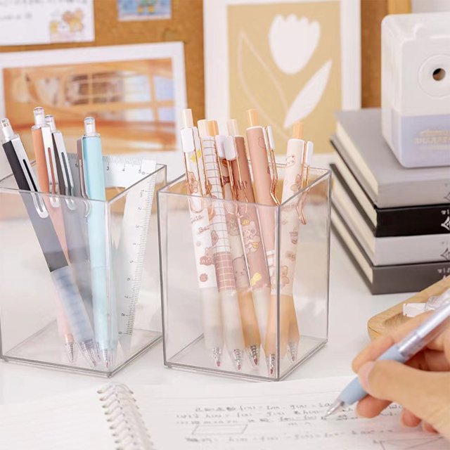 Nilvkv Porte-Stylo à Grande Capacité, Baril de Papeterie Transparent pour l'École et la Maison. l'Image Montre Deux Porte-Stylos en Acrylique Transparent, Chacun Contenant une Variété de Stylos et d'Articles de Papeterie. les Porte-Stylos Sont Placés sur un Bureau à Côté d'Un Cahier, d'Une Écriture Manuelle avec un Stylo, et d'Autres Accessoires de Bureau comme des Livres et un Taille-Crayon. les Porte-Stylos au design Moderne Offrent un Grand Espace de Rangement pour Organiser des Stylos, des Règles et d'Autres Petits Outils,