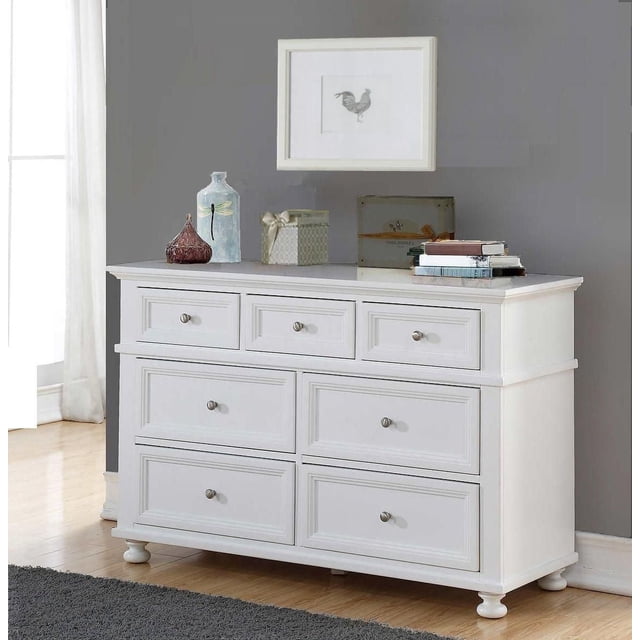 Concord Baby Classic Dresser Changer in white with seven drawers featuring silver metal knobs and decorative molding, standing on rounded feet on a wooden floor against a gray wall, topped with decorative vases, a gift box, and stacked books beneath a framed rooster print in a nursery.