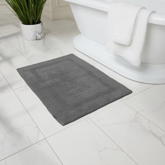Fabstyles Basix Soft & Absorbent Reversible Cotton Bath Rug in grey placed on white marble tile floor beside a white freestanding bathtub with a white towel draped over the side and a green plant in a marble pot nearby, ideal for shower, tub, or sink areas.