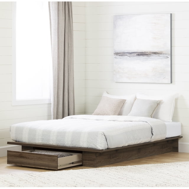 South Shore Tao Platform Bed with Storage Drawer for Adult in natural walnut finish, shown in a bright bedroom with white shiplap walls and light hardwood floors. The low-profile platform bed features a large hidden drawer with metal slides and a cut-out handle, partially open to reveal storage space ideal for linens or clothes. The bed is dressed with light grey bedding and neutral pillows, positioned near a window with beige curtains and a neutral abstract painting on the wall.