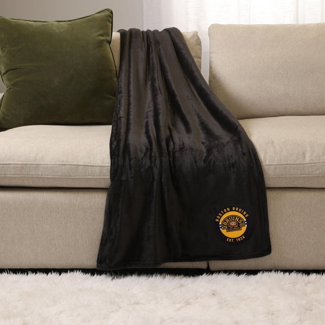 NHL, NBA, MLB Black Sports Throw Blanket, 50" x 60", draped over a beige sofa with a green pillow, featuring a Boston Bruins logo patch in yellow and black on the lower corner, made of soft plush polyester suitable for indoor and outdoor use.