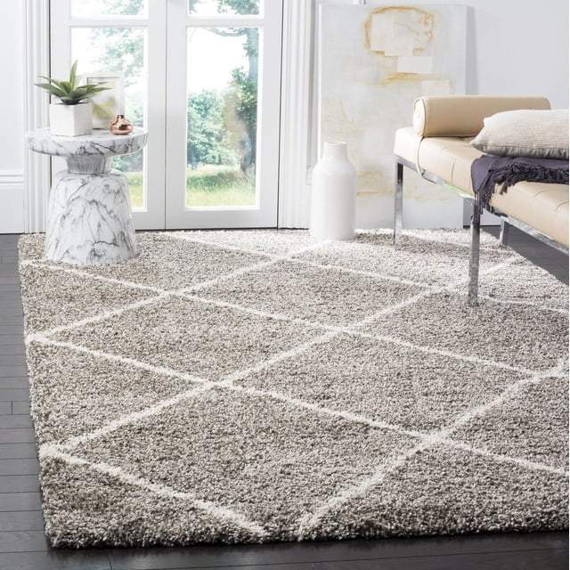 Safavieh Hudson Amias Geometric Shag Area Rug featuring a thick plush pile with ivory diamond tile patterns on a taupe background, set on dark hardwood floors in a bright room with glass doors, styled with a beige leather bench, marble side table with plant, and white ceramic vase, suitable for enhancing texture in urban or modern décor.