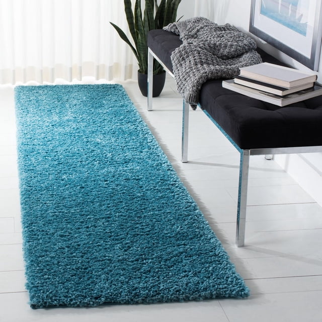 SAFAVIEH August Carlene Solid Plush Shag Area Rug in blue, a 2'3" x 8' rectangular runner with a thick polypropylene plush pile, placed on white hardwood flooring beside a black bench with chrome legs, featuring a gray knitted throw, stacked books, and a tall green plant near sheer white curtains, suitable for adding softness and style to hallways or entryways.