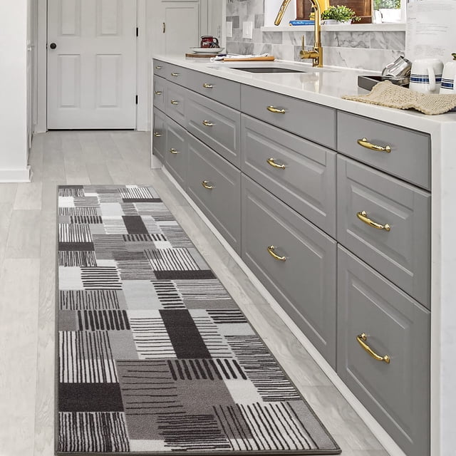 BY COCOON 2'x8' runner rug in grey displayed in a modern kitchen setting. The washable polypropylene rug features a geometric pattern with varying shades of grey and black. Its low pile design ensures functionality in high-traffic areas, such as hallways or kitchens, while the non-slip backing provides safety. Positioned along a row of grey cabinets with gold handles, the rug complements the sleek decor and is easy to clean.