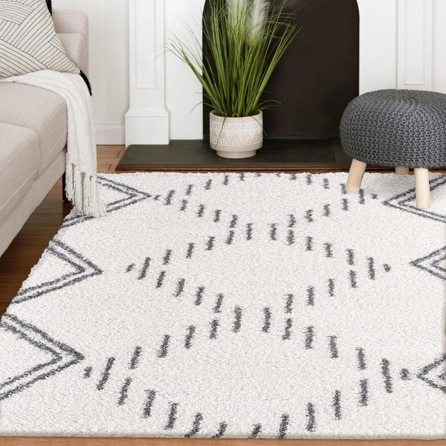 Andela Cream and Grey Woven Area Rug featuring large charcoal diamond patterns on an ivory background, displayed in a living room with a beige sofa, white throw, grey knitted pouf with wooden legs, and a potted green plant near a black fireplace hearth.