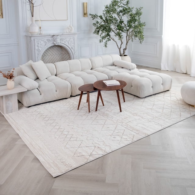 Rug Branch Astra Boho Indoor Area Rug in beige cream with abstract geometric patterns, on light wood herringbone floor in a bright living room with a tufted cream sectional sofa, two round wooden nested coffee tables with a book, white marble fireplace, and large potted green plant near sheer white curtains.