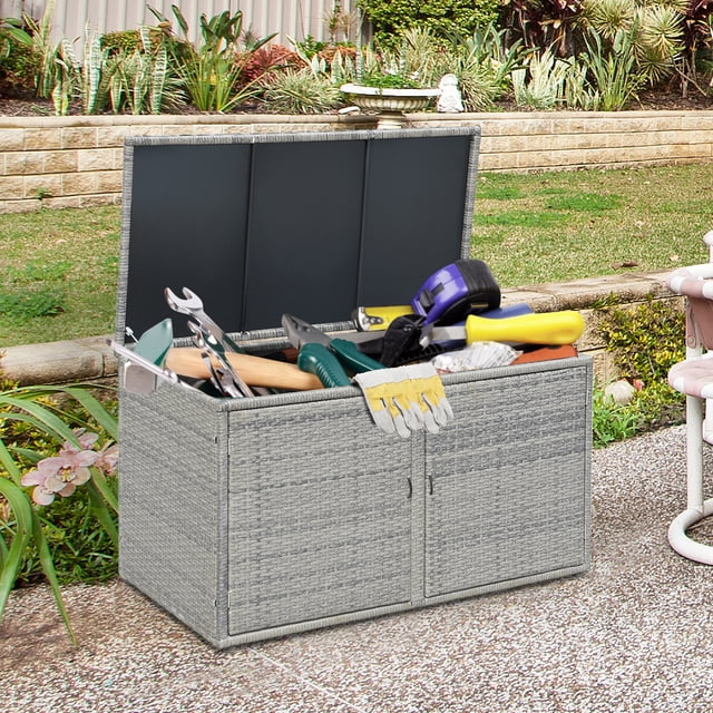 Gymax Coffre de Rangement en Rotin de 88 Gallons pour Terrasse Extérieure avec Tablette et Porte en Gris, Montré dans un Décor de Jardin Extérieur. le Coffre Présente un Cadre en Acier durable avec une construction en Rotin Serrée et un design à Deux Couches pour un Grand Rangement d'Outils, de Coussins et d'Autres Articles. le Couvercle Supérieur Est Ouvert, Révélant Divers Outils de Jardin à l'Intérieur. le Coffre Comprend Aussi des Portes avant avec Poignées pour un Accès Facile et des Pieds Ajustables pour la Stabilité sur les surfaces Irrégulières.