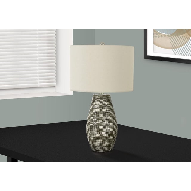 Monarch Specialties Lighting 24"h table lamp with a grey resin vase-shaped base featuring raised dotted texture, ivory fabric drum shade, and silver metal finial, placed on a black table near a window with white blinds and grey walls, suitable for living rooms or bedrooms.