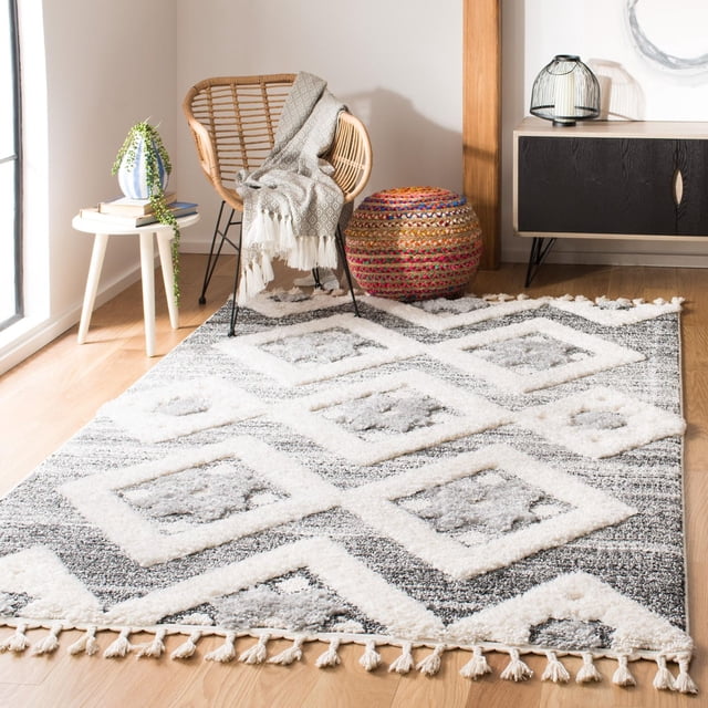 Safavieh Moroccan Tassel Shag Dawson Moroccan Geometric Area Rug with plush grey and white shag texture and tassel fringe on light wood flooring, styled with a rattan chair draped with a grey patterned throw, a colorful woven pouf, a white side table with books and a trailing plant, and a black wood cabinet with a decorative lantern, suitable for enhancing comfort and style in accent rug settings.