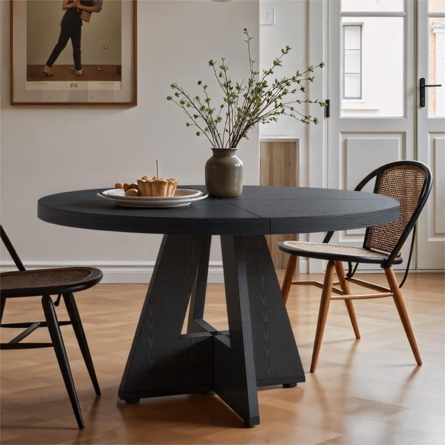 Fnova 47" Coffee Table, a round dining table for 4 people, shown in black with a modern farmhouse design. The table features a 1.18-inch thick waterproof and scratch-resistant wooden top and 1.57-inch thick legs with a stable structure. It is styled in a bright dining room with wooden flooring, paired with chairs (not included) and a decorative vase with branches on top.