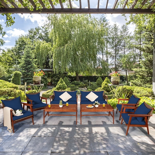 Costway 8PCS Patio Rattan Furniture Set with acacia wood frame and navy cushions, displayed in an outdoor garden setting. The set includes four single sofas, two loveseats, and two coffee tables, all featuring hand-woven rattan and thick, removable cushions for comfort. The furniture is arranged on a stone patio under a pergola, surrounded by lush greenery and decorative planters.