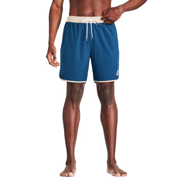 Reebok Short de Planche pour Homme à Logo Uni de 7 Pouces en Bleu avec Taille Élastique beige et Cordon de Serrage Blanc, avec un Logo Reebok Blanc sur la Jambe Gauche, Séchage Rapide et Résistant au Chlore et à l'Eau de Mer, Convient à la Natation Ou aux Vêtements de Plage, Inspiré d'Un Homme Debout Pieds Nus sur un Fond Blanc.