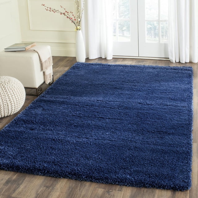 Safavieh Milan Harlow Solid Shag Area Rug in navy blue, a rectangular plush synthetic yarn shag rug on wood flooring, displayed in a bright room with white curtains, a cream ottoman with a throw, and a knitted pouf, suitable for adding warmth and texture to contemporary home décor.