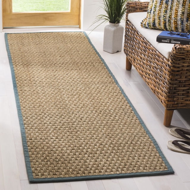Safavieh Natural Fiber Arbor Border Area Rug featuring woven sisal texture in natural beige with a muted blue fabric border, placed on light hardwood flooring in a hallway beside a wicker armchair and potted plant, suitable for coastal or casual interiors and often used as a durable runner rug for home or office.