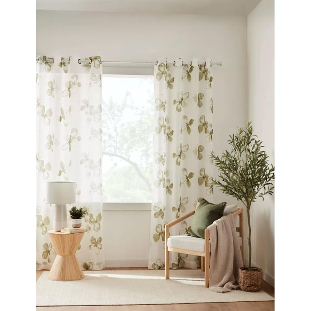 Bouclair Olive Nima Rideau Transparent 52x84 Po (132x213 cm) Exposé dans une Pièce Lumineuse et Moderne. le Rideau Présente un Délicat Motif floral en Vert Olive sur un Tissu polyester Transparent, Permettant à la Lumière Naturelle de Filtrer à Travers. Il Est Suspendu sur une Tringle à Rideaux Argentée avec Oeillets, Encadrant une Fenêtre avec une Vue de Verdure. le Cadre Comprend une Chaise en Bois avec un Coussin Vert et un Jeté beige, une Petite table d'Appoint en Bois avec une Lampe et des Plantes en Pot, et un Panier Tressé avec un Arbre