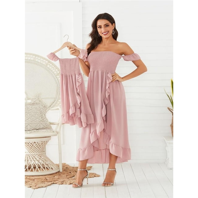 Mother Daughter Dress Ruffle Family Matching Dresses Off Shoulder Mom And Daughter Matching Clothes Summer Mommy And Me Clothes. The image shows a woman wearing an off-shoulder ruffle dress in a soft pink color, paired with a matching smaller dress for a child displayed on a hanger. The dresses feature a smocked bodice and cascading ruffle details, ideal for summer outings. The setting is a bright, minimalistic room with white walls and wicker furniture.