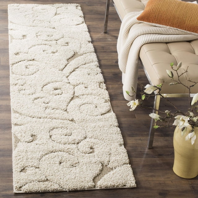 SAFAVIEH Florida Tapis Shag à Vignes Florales Douglas en Ton Crème avec Motifs de Vignes Damassées Sculptées Asymétriques, Fabriqué à Partir de Fils Synthétiques Tissés Mécaniquement Durables, Exposé sur Plancher de Bois Franc Foncé à Côté d'Un Banc en Cuir beige avec Jeté Tricoté et Coussin Couleur Rouille, Près d'Un vase Jaune avec des Fleurs Blanches.