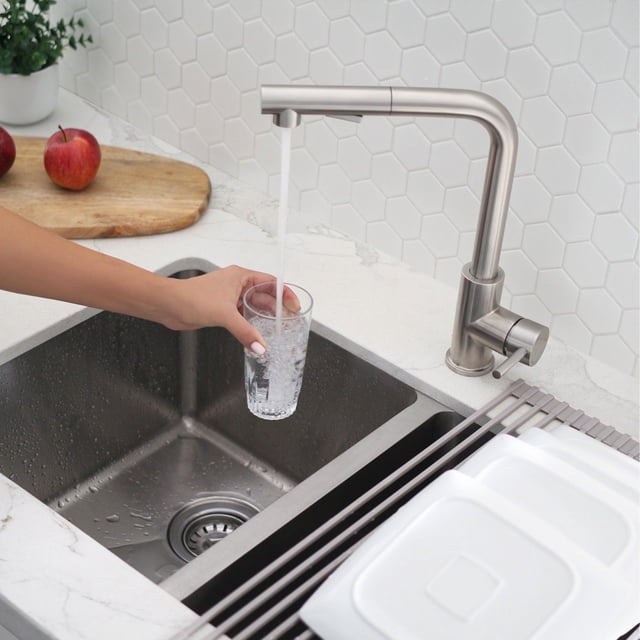 Robinet de Cuisine Élégant de 12 5⁄8 Pouces Ht à une Poignée en Acier Inoxydable Brossé à Bec Rétractable K-130S Installé dans une Cuisine Moderne. le Robinet a une Finition en Acier Inoxydable Brossé, une Poignée Unique pour un Contrôle Précis, et un Vaporisateur à Bec Rétractable avec Deux modes pour Jet et Vaporisation d'Eau. une Main Remplit un Verre d'Eau, Démontrant Sa Fonctionnalité. la Zone de l'Évier Présente une Bassine en Acier Inoxydable, un Comptoir Blanc et un Dosseret hexagonal en Carreaux.