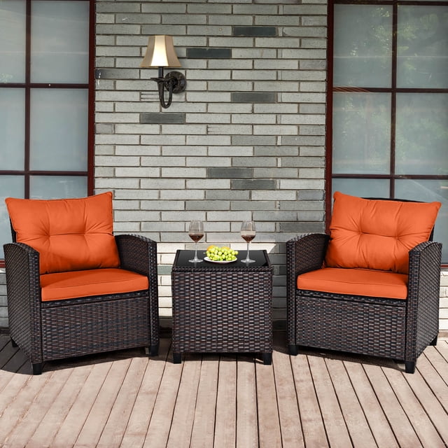 Costway 3 PCS Patio Rattan Furniture Set Cushioned Conversation Set Sofa Orange. This set includes two mix-brown rattan armchairs with orange cushions and a square coffee table with a tempered glass top. The cushions are thick, removable, and feature zippered covers for easy cleaning. Designed for outdoor use, the set is shown on a wooden patio with a brick wall backdrop, suitable for patios, gardens, or poolside areas.