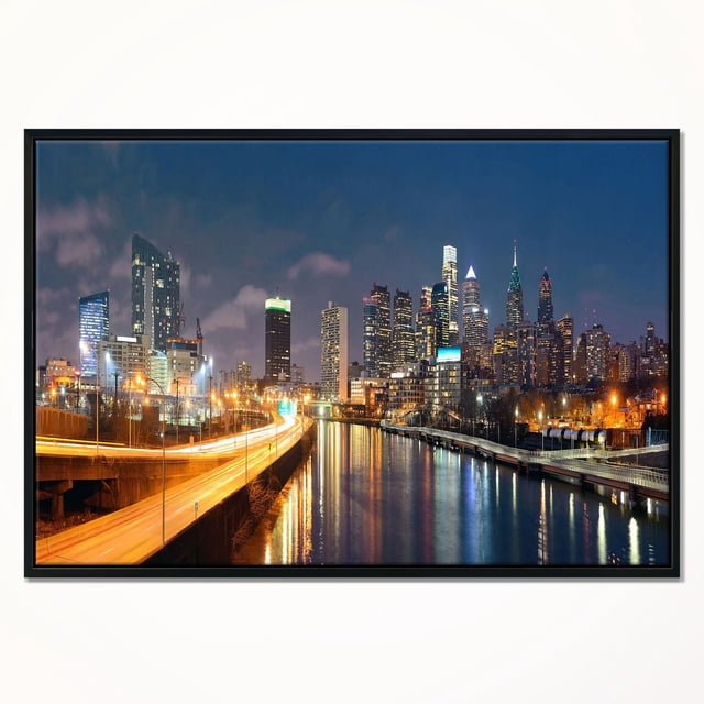 Design Art Philadelphia Skyline at Night Framed Canvas Art Print featuring a centered front view of Philadelphia’s illuminated skyline at night with skyscrapers reflecting on the river and a lit highway, printed on fade-resistant canvas stretched over a 1-inch wood subframe and gallery-wrapped with a black floating frame, including hanging kit for easy installation.