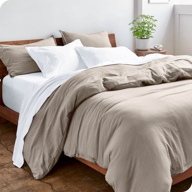 Bare Home Housse de Couette Sablée - Ultra Douce - Premium 1800 Collection - Ensemble 3 Pcs - Très Grand, Galet. l'Image Montre une Housse de Couette Couleur Galet avec une texture Douce et Sablée et des Taies d'Oreiller Assorties sur un Cadre de Lit en Bois. la Literie Est Conçue dans une Chambre Moderne avec une Plante en Pot sur une table d'Appoint, Soulignant Son Attrait chic et Confortable.