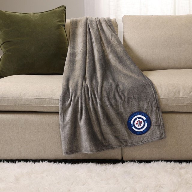 NHL, NBA, MLB Grey Sports Throw Blanket, 50" x 60", draped over a beige sofa with a green cushion, made of soft plush polyester featuring a Winnipeg Jets logo patch in blue, white, and red on the lower corner, suitable for indoor use and cozy warmth.