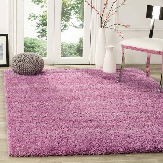 Safavieh California Solid Plush Shag Area Rug in soft pink, displayed on light wood flooring in a bright room with a gray knit pouf, white accent chair with chrome legs, and two white vases with flowering branches near glass doors overlooking greenery, ideal for adding plush comfort to living spaces.