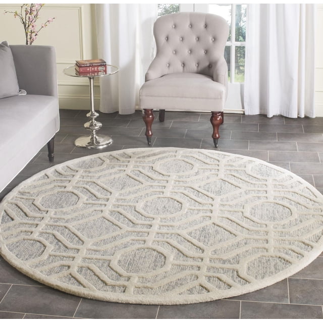 Safavieh Cambridge Johan Geometric Area Rug, round with a textured geometric pattern in cream and light gray wool, placed on a dark gray tiled floor in a living room with a light gray upholstered armchair with wooden legs and a small round silver side table holding books.