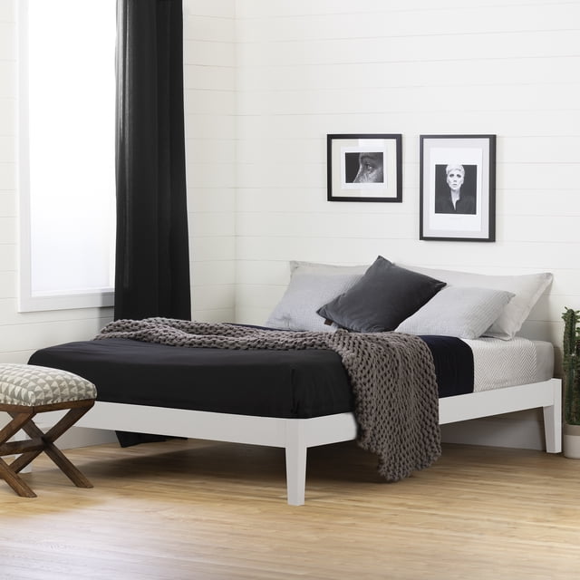 South Shore Vito Queen Solid Wood Platform Bed in white with a minimalist wooden frame and angled legs, dressed in black and gray bedding with a chunky knit throw. The bed is placed in a room with white shiplap walls, light wood flooring, two black-framed monochrome photos above, and a patterned cushioned stool nearby.