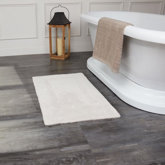 Fabstyles Basix Tapis de Bain en Coton Réversible Doux et Absorbant Blanc, Placé sur un Plancher de Salle de Bain Aspect Bois Foncé, à Côté d'Une Baignoire Autoportante Blanche avec une Serviette beige Drapée sur le Bord et une Lanterne en Bois avec une Bougie en Arrière-Plan, Fabriqué à Partir de 100 % de Coton Bouclé pour l'Absorption et la Durabilité, Convient comme Tapis de Douche Ou de Baignoire.