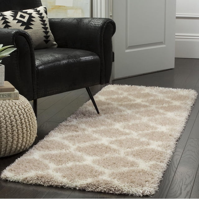Safavieh Montreal Lennart Geometric Shag Area Rug in beige and cream with a plush diamond pattern, placed on dark hardwood flooring beside a black textured armchair with a black and white geometric pillow, suitable for contemporary or transitional rooms.